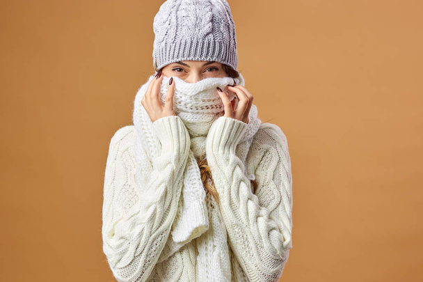 Nice girl dressed in white knitted sweater and hat closes her face with white scarf on a beige background in the studio - 写真・画像