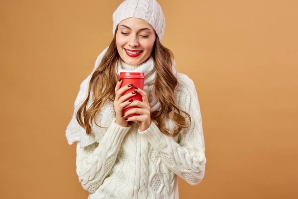 Smiling girl dressed in white knitted sweater and hat holds a red cup in her hands on a beige background in the studio - 写真・画像