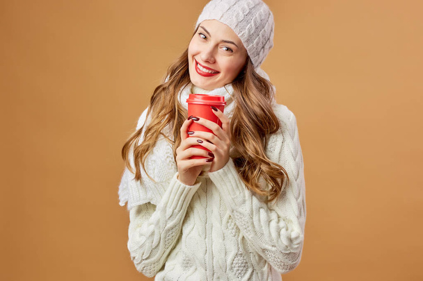 Smiling girl dressed in white knitted sweater and hat holds a red cup in her hands on a beige background in the studio - Foto, Bild