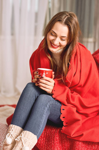 Girl dressed in white knitted sweater and jeans covered in red wrap is sitting with a red cup of hot drink in her hands - Foto, Imagen