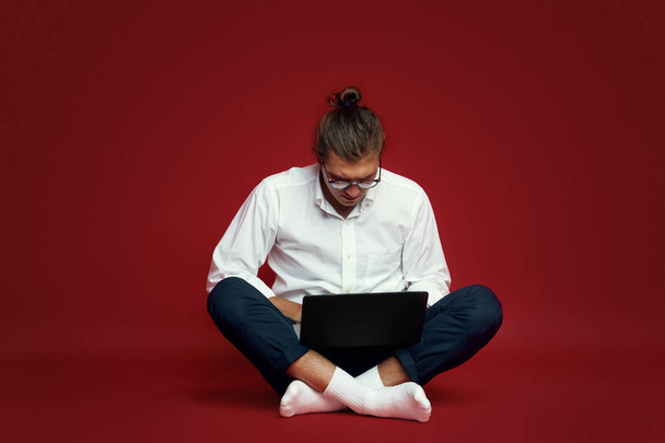 Πορτρέτο του σοβαρή νεαρός άνδρας στο λευκό πουκάμισο ψάχνει στο φορητό υπολογιστή, ενώ κάθεται σε ένα πάτωμα που απομονώνονται σε κόκκινο φόντο - Φωτογραφία, εικόνα
