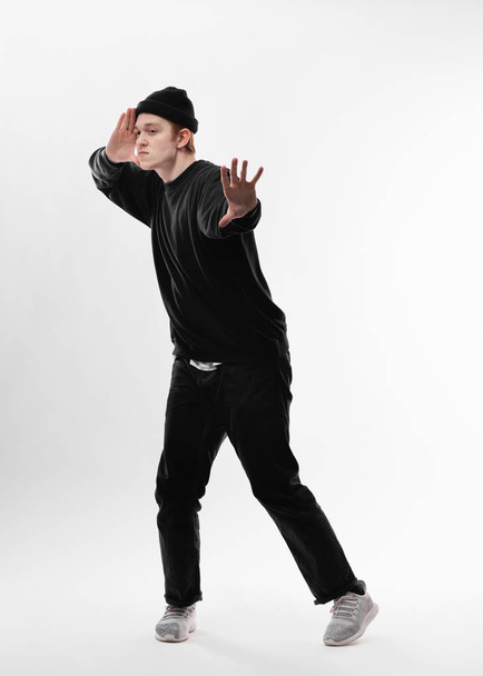 Jonge freestyle danser, gekleed in zwarte jeans, trui, hoed en grijze sneakers is dansen in de studio op de witte achtergrond - Foto, afbeelding