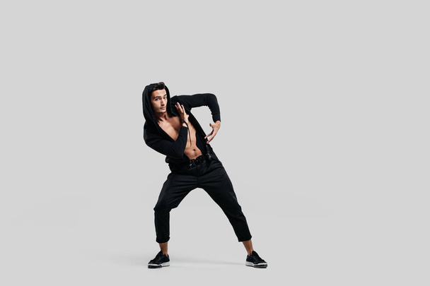 Young handsome young dancer dressed in black pants, a sweatshirt on a naked torso is dancing street dance. He makes stylized movements with his hands - Foto, imagen