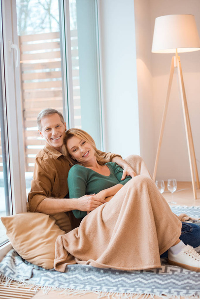 счастливый муж обнимает жену сидя вместе на полу у большого окна
  - Фото, изображение