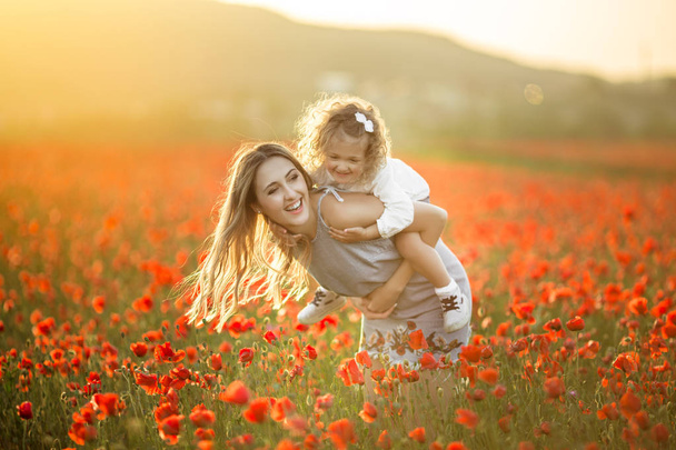 Beautiful child girl with young mother are having fun in field of poppy flowers over sunset lights - Foto, Imagen