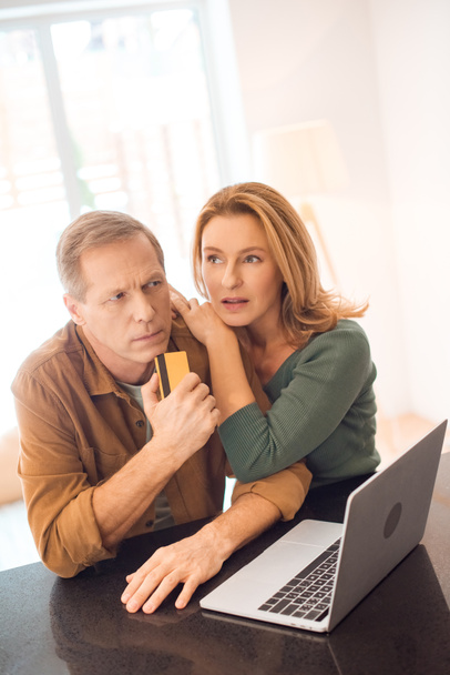 thoughtful couple standing by laptop while husband holding credit card - Photo, Image