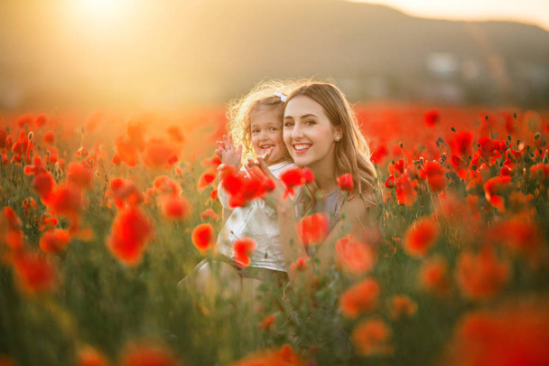 Beautiful smiling child girl with young mother are having fun in field of poppy flowers over sunset lights - Foto, afbeelding