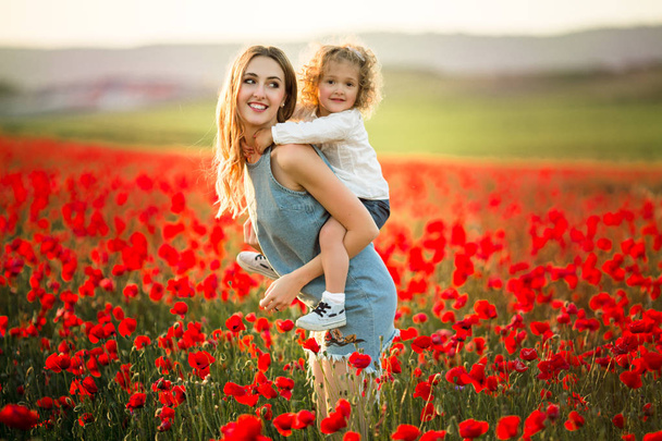 Beautiful smiling child girl with young mother are having fun in field of poppy flowers over sunset lights - Photo, image