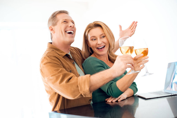 ラップトップ テーブルで立ちながら白ワインのガラスをチリンと興奮のカップル - 写真・画像