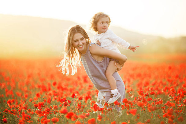 Beautiful smiling child girl with mother are having fun in field of red poppy flowers over sunset lights - Photo, Image