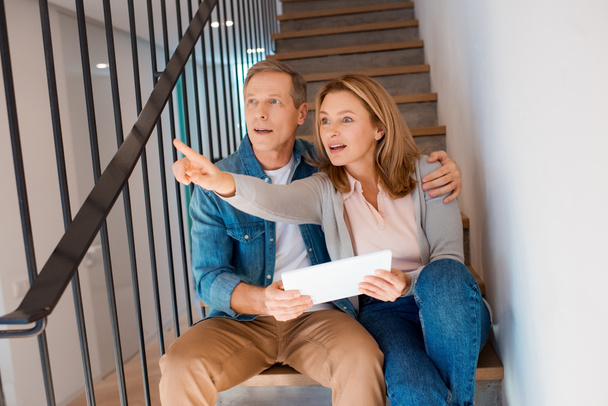 woman pointing with finger while sitting on stairs with husband and using digital tablet - Photo, image