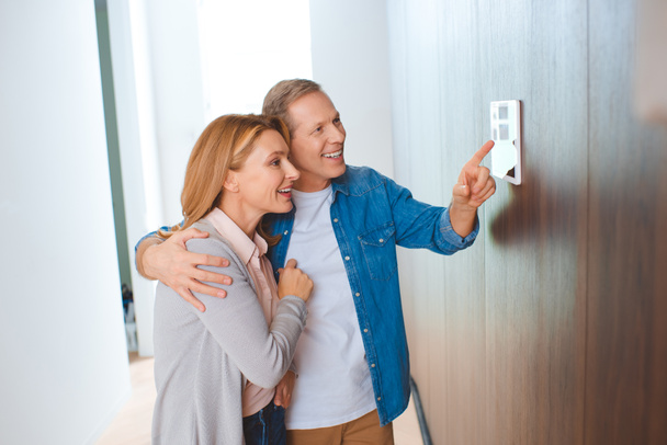 ευτυχισμένος άνθρωπος δείχνει τον πίνακα ελέγχου smart home ενώ αγκαλιάζει σύζυγος - Φωτογραφία, εικόνα