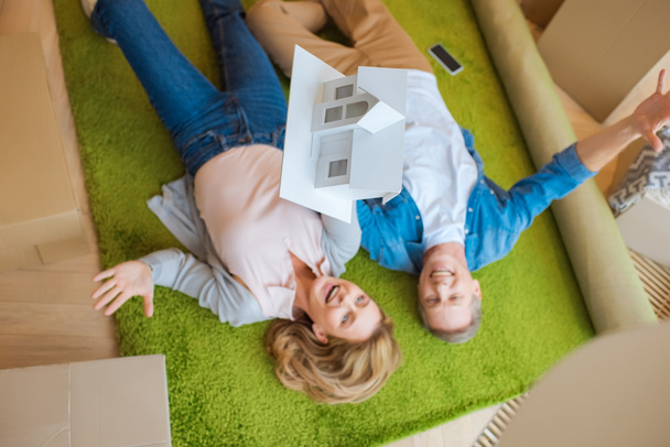 overhead view of happy couple lying on green carpet with house model - Fotó, kép