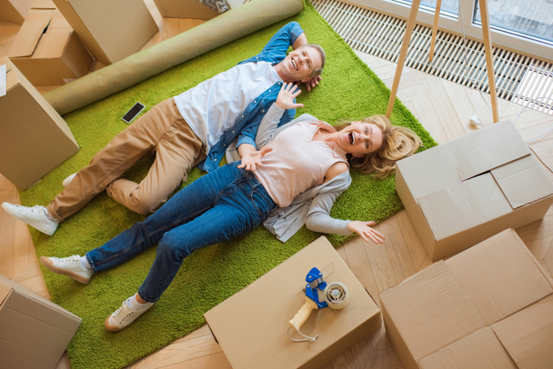 happy couple lying on floor on green carpet surrounded with cardboard boxes - Fotografie, Obrázek