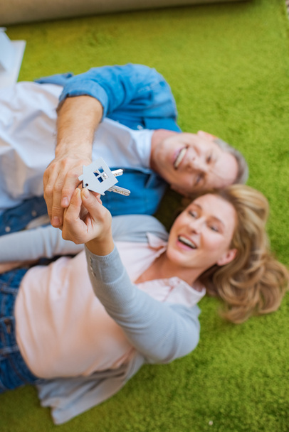 enfoque selectivo de pareja feliz sosteniendo llaves con baratija modelo de casa mientras está acostado en la alfombra verde
  - Foto, imagen