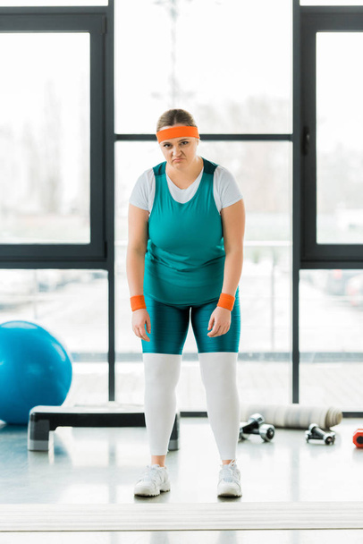 засмучена надмірна вага жінка стоїть в спортивному одязі в спортзалі
 - Фото, зображення