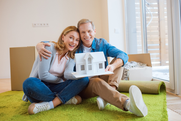 happy couple hugging and demonstrating house model while sitting on floor on green carpet - Photo, Image