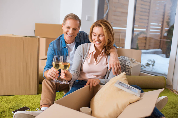 happy couple clinking glasses of white wine while sitting by cardboard boxes - Φωτογραφία, εικόνα