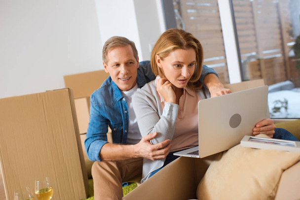 couple en utilisant un ordinateur portable tout en étant assis près des boîtes en carton à la nouvelle maison
 - Photo, image