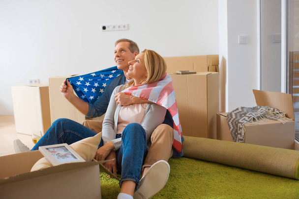 happy couple wrapped in usa national flag looking up while sitting near cardboard boxes at new home - 写真・画像