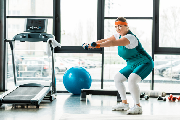 cheerful plus size woman exercising with dumbbells near treadmill  - Zdjęcie, obraz