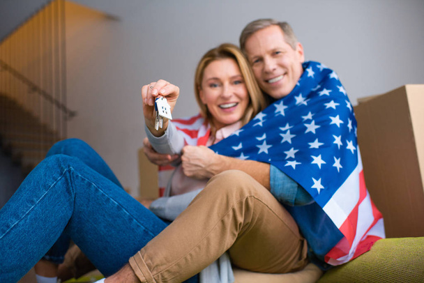 selektiver Fokus der Frau, die Schlüssel mit Hausmodell Schmuckstück hält, während sie mit ihrem Mann die US-Nationalflagge einwickelt - Foto, Bild