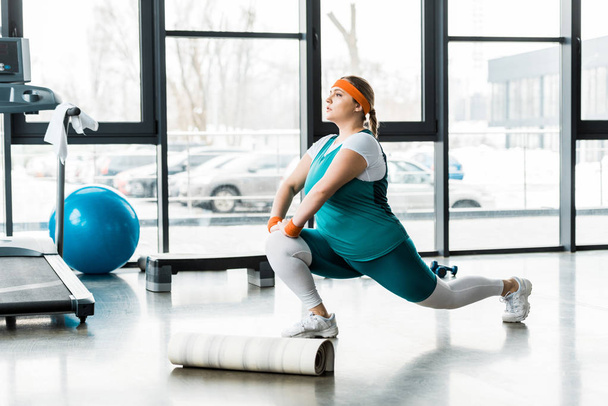 plus size woman in sportswear stretching near fitness mat and treadmill  - Fotó, kép