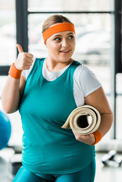 Veselá obézní žena ukazuje palcem podržíte fitness mat v tělocvičně - Fotografie, Obrázek