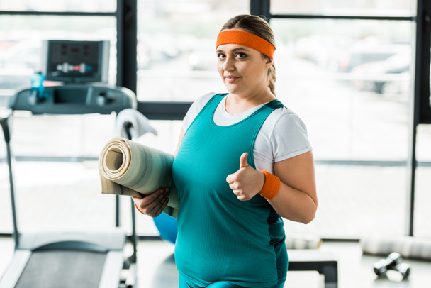 happy overweight woman showing thumb up while holding fitness mat in gym - Fotó, kép