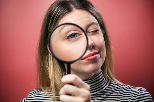 Cheerful young woman looking through magnifying glass at the camera over pink background. - Photo, Image