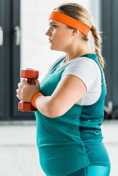 vista lateral de la chica con sobrepeso grave sosteniendo la mancuerna en el gimnasio
 - Foto, Imagen