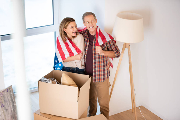 Selektiver Fokus eines glücklichen Paares, das sich in eine US-Nationalflagge hüllt, während es in der Nähe von Pappkartons im neuen Zuhause steht - Foto, Bild
