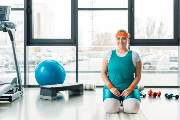χαρούμενα συν μέγεθος γυναίκα που κάθεται στο γυμναστήριο mat κοντά σε αθλητικό εξοπλισμό - Φωτογραφία, εικόνα