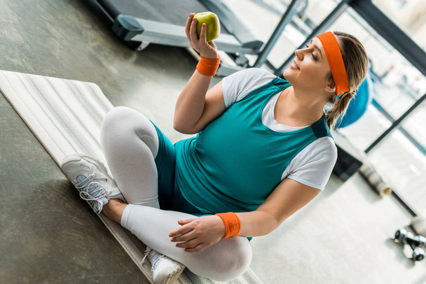 plus size woman sitting on fitness mat with crossed legs and looking at apple - Foto, imagen