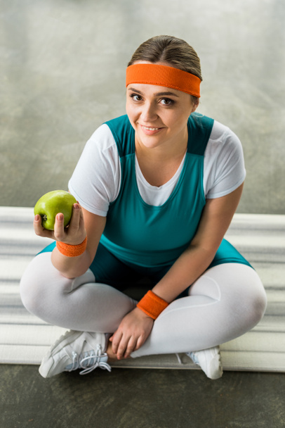 bovenaanzicht van happy plus grootte meisje zittend op fitness mat met gekruiste benen en lekkere appel te houden - Foto, afbeelding