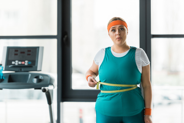 surprised plus size woman measuring waist in gym - Photo, Image