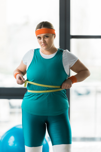 dissatisfied overweight girl measuring waist in gym - Foto, Imagen