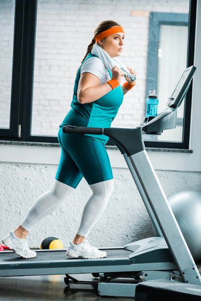 tired overweight girl with towel on shoulders running on treadmill in gym - Foto, Imagem