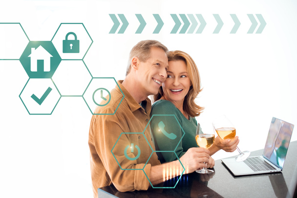 happy couple holding glasses of white wine while standing by table with laptop, smart home concept - Foto, imagen
