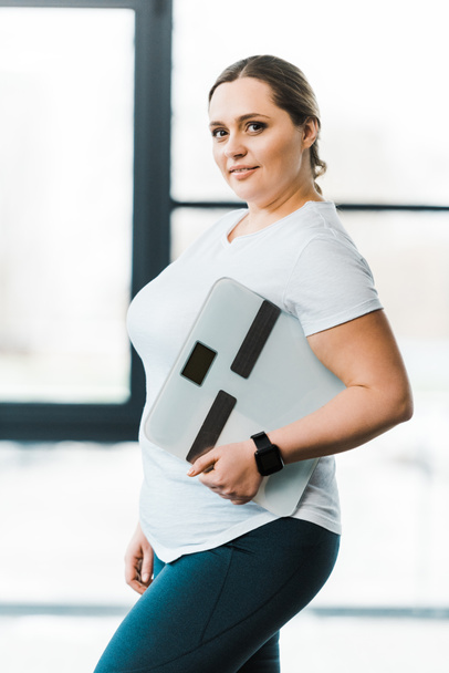 веселая женщина с избыточным весом улыбается, держа весы
 - Фото, изображение
