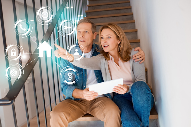 Frau zeigt Hand, während sie mit Mann auf Treppe sitzt und digitales Tablet nutzt, Smart-Home-Konzept - Foto, Bild