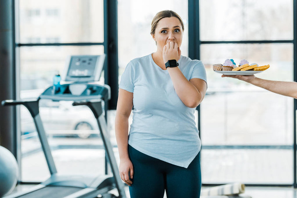 levágott képe ember tartja közelében száj edzőteremben, amely túlsúlyos nő finom péksütemények lemez - Fotó, kép