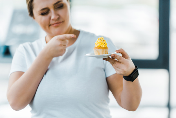 cheerful overweight woman pointing with finger at tasty cupcake in gym - Fotó, kép
