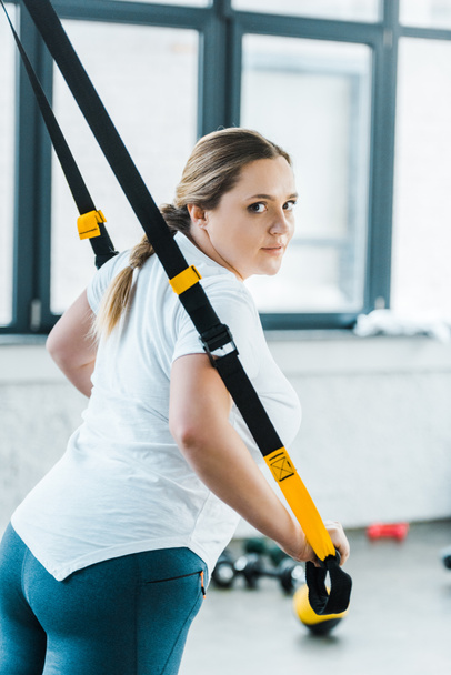 attractive overweight woman training arms with suspension straps in gym - Photo, Image