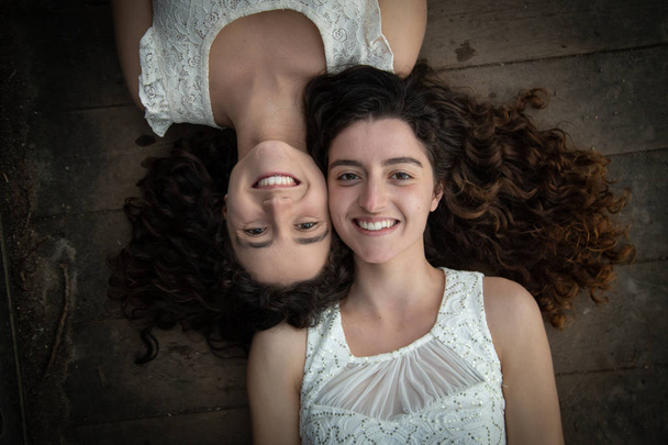 From above of young and beautiful twin sisters in white dresses lying opposite on wooden floor smiling at camera - Zdjęcie, obraz