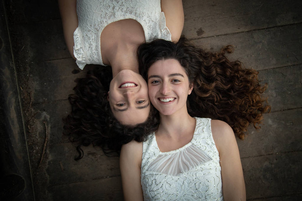 From above of young and beautiful twin sisters in white dresses lying opposite on wooden floor smiling at camera - Photo, Image