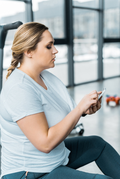 chica con sobrepeso usando teléfono inteligente mientras está sentado en el gimnasio
 - Foto, Imagen