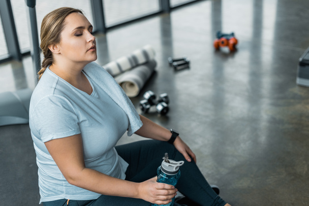 unavený plus velikosti žena odpočívá se zavřenýma očima a drží láhev s vodou - Fotografie, Obrázek