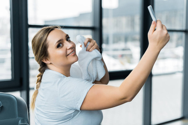 happy overweight girl taking selfie while holding towel in gym - Photo, Image