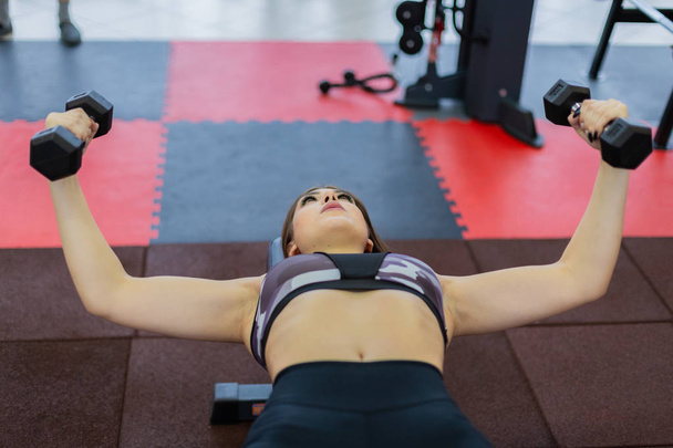 Красивая девушка тренирует грудные мышцы лежа гантели на скамейке в тренажерном зале
. - Фото, изображение